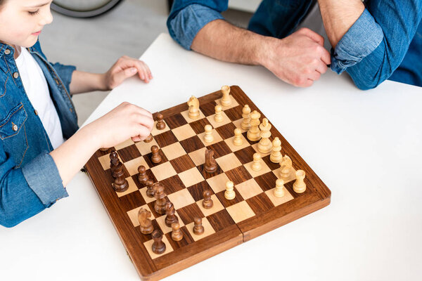 cropped view of father and son in denim sitting at table and playing chess at home