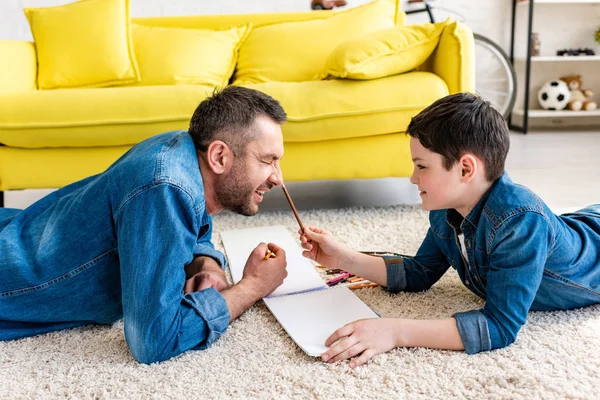 リビングルームで色鉛筆でカーペットの上に横たわっている父と息子 — ストック写真