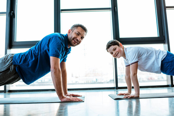 happy father and son looking at camera while doing push up exercise at gym