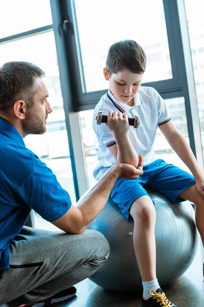 スポーツセンターでダンベルで運動するフィットネスマットで息子を助ける父親 — ストック写真