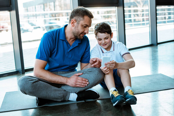 father and son sitting on fitness mat and using smartphone at gym