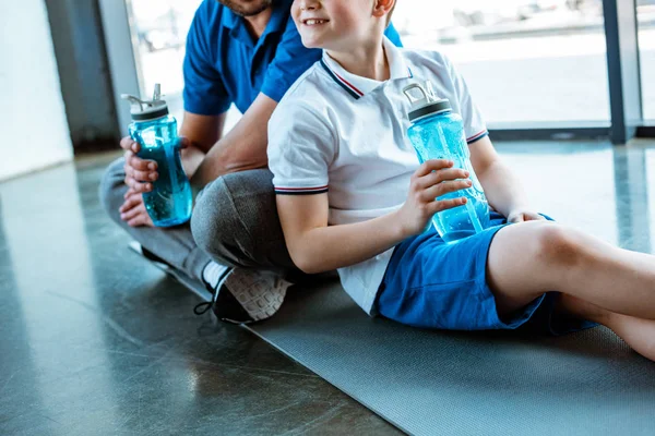 cropped view of father and son sitting on fitness mat with sport bottles at gym