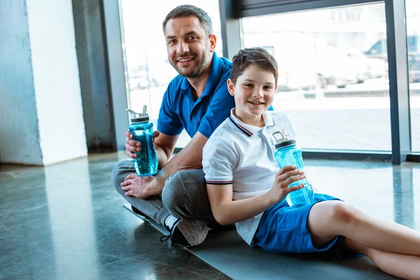 father and son sitting on fitness mat with sport bottles and looking at camera at gym