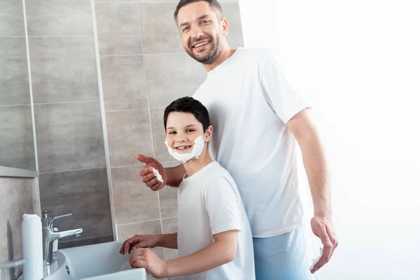 バスルームで顔にシェービングクリームを持つ息子の近くに笑顔の父 — ストック写真