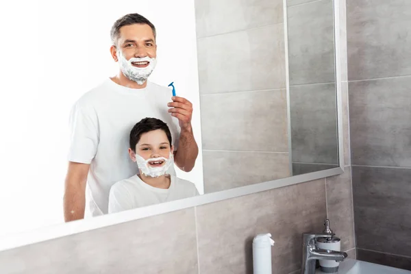 smiling son and father with shaving cream on faces looking at camera in bathroom