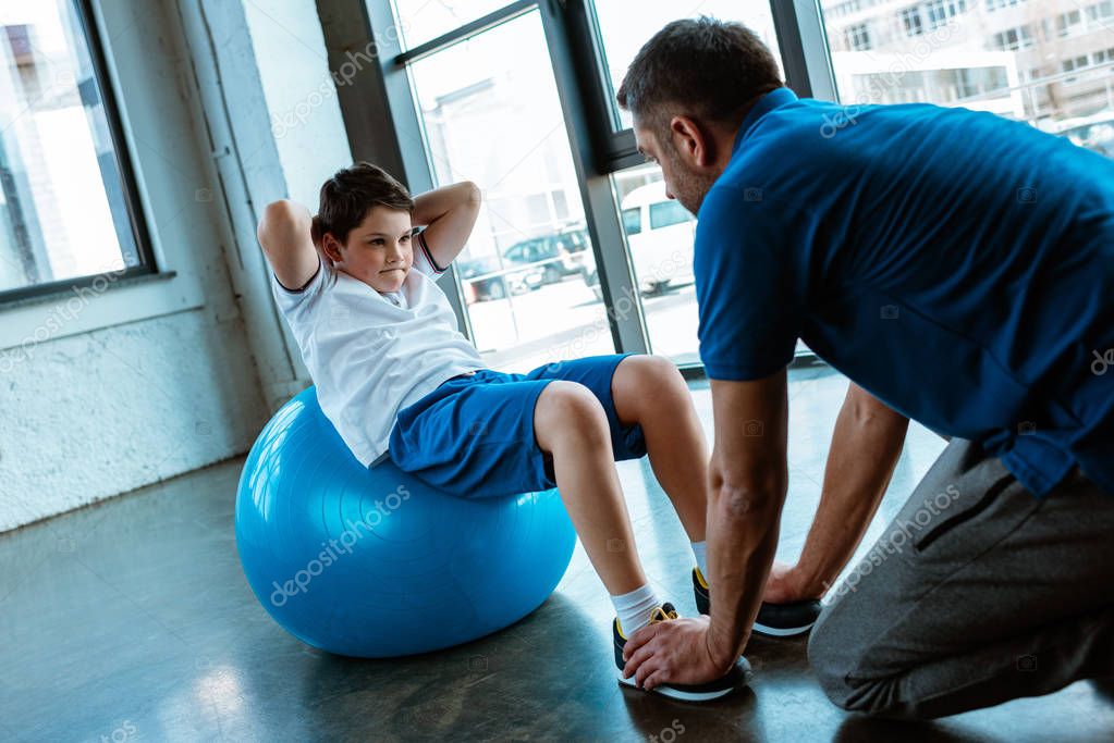 father helping son sitting on fitness ball and doing sit up exercise at gym