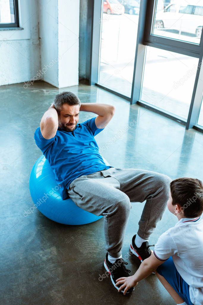 son helping father sitting on fitness ball and doing sit up exercise at gym