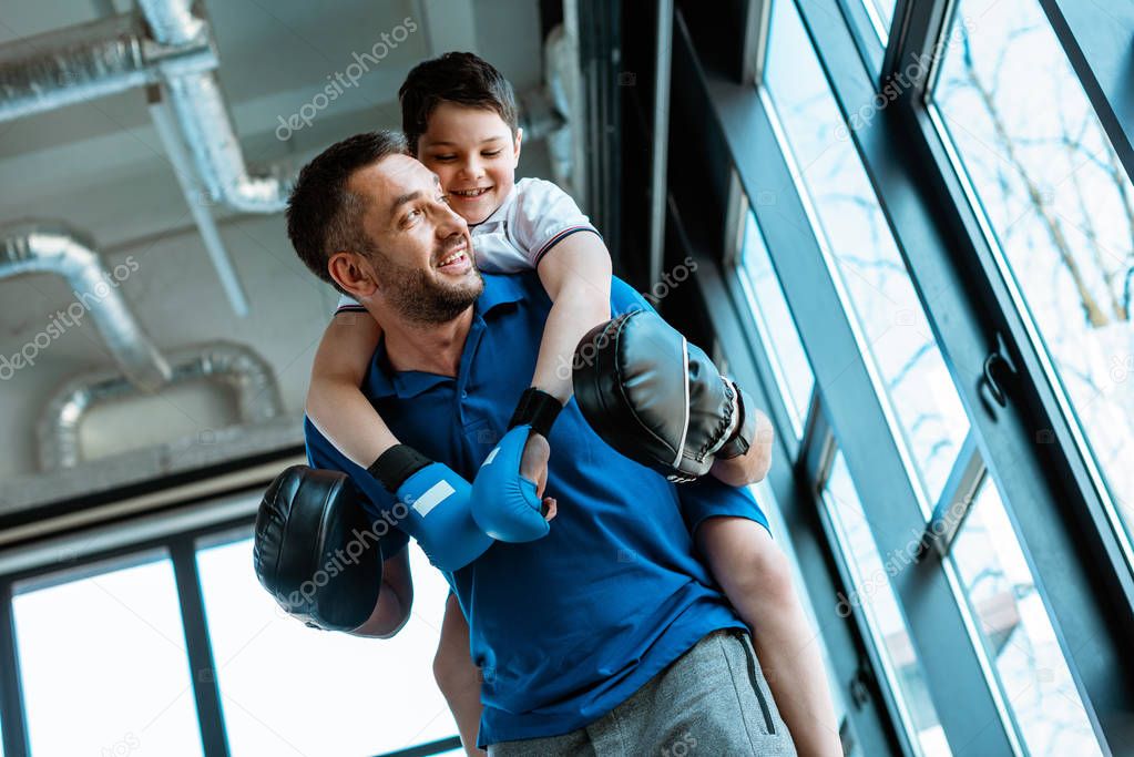 father giving piggyback ride to smiling son in boxing gloves at gym