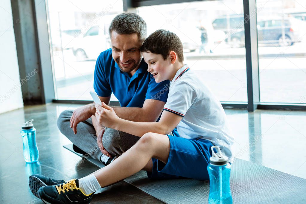 father and son sitting on fitness mat and using smartphone at gym