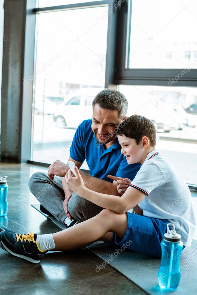 smiling father and son sitting on fitness mat and using smartphone at gym