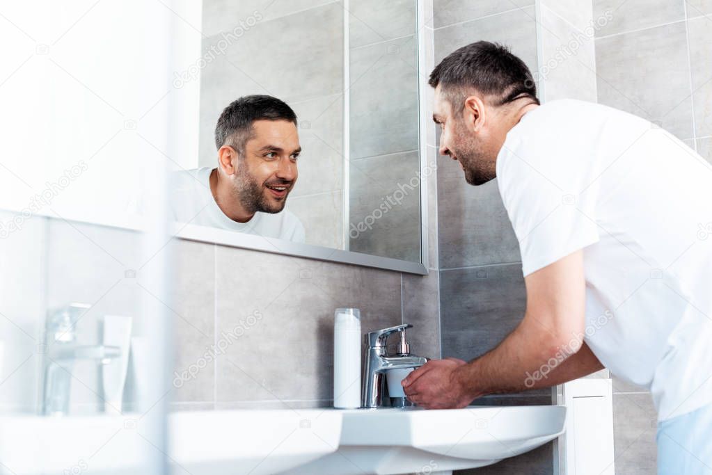 handsome smiling man looking in mirror and washing hands in bathroom during morning routine