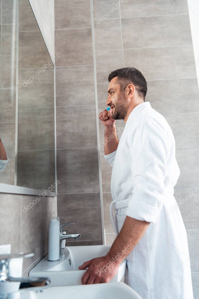 handsome man in white bathrobe brushing teeth in bathroom during morning routine