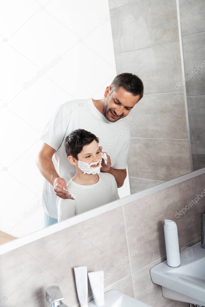 smiling father applying shaving cream on face of son in bathroom