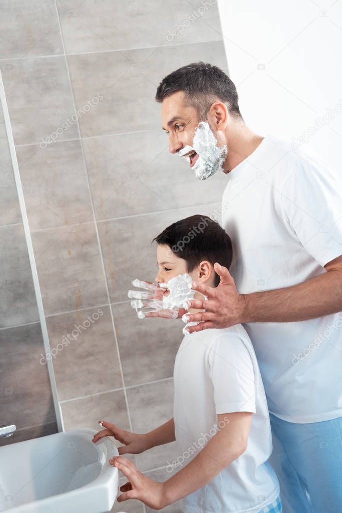 smiling father applying shaving cream on face of son in bathroom in morning
