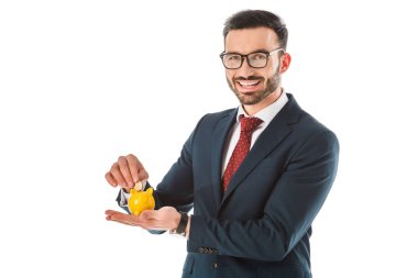 cheerful businessman putting coin into piggy bank and looking at camera isolated on white clipart