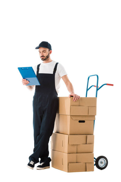 serious delivery man standing near hand truck loaded with cardboard boxes and looking at clipboard isolated on white