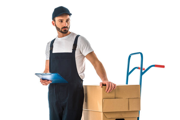 thoughtful delivery man holding clipboard while standing near hand truck loaded with carton boxes isolated on white
