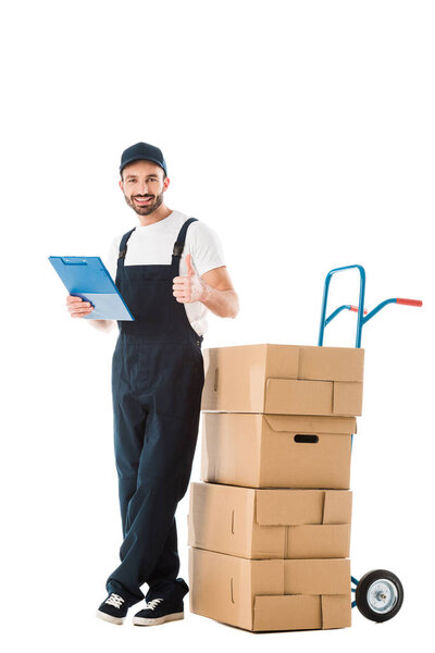 happy delivery man standing near hand truck with carton boxes and showing thumb up isolated on white