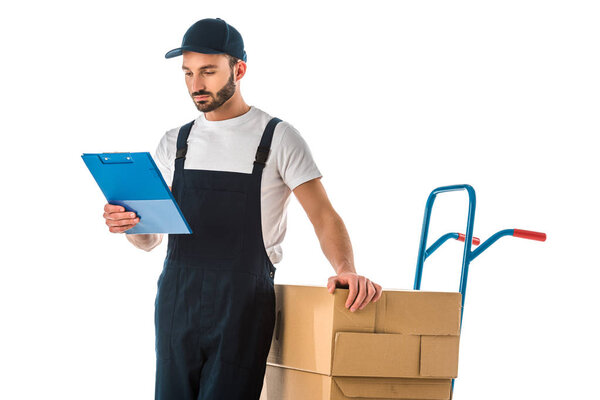 serious delivery man looking at clipboard while standing near hand truck loaded with cardboard boxes isolated on white
