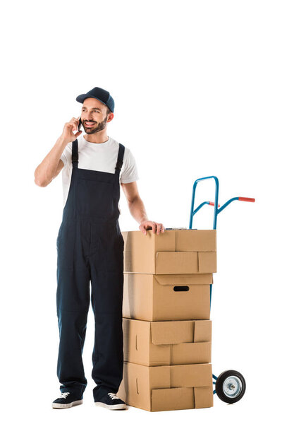cheerful delivery man talking on smartphone while standing near hand truck with carton boxes isolated on white