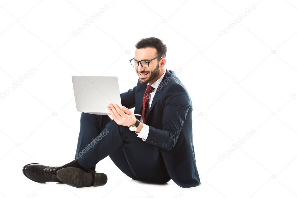 smiling businessman in glasses using laptop while sitting on white background