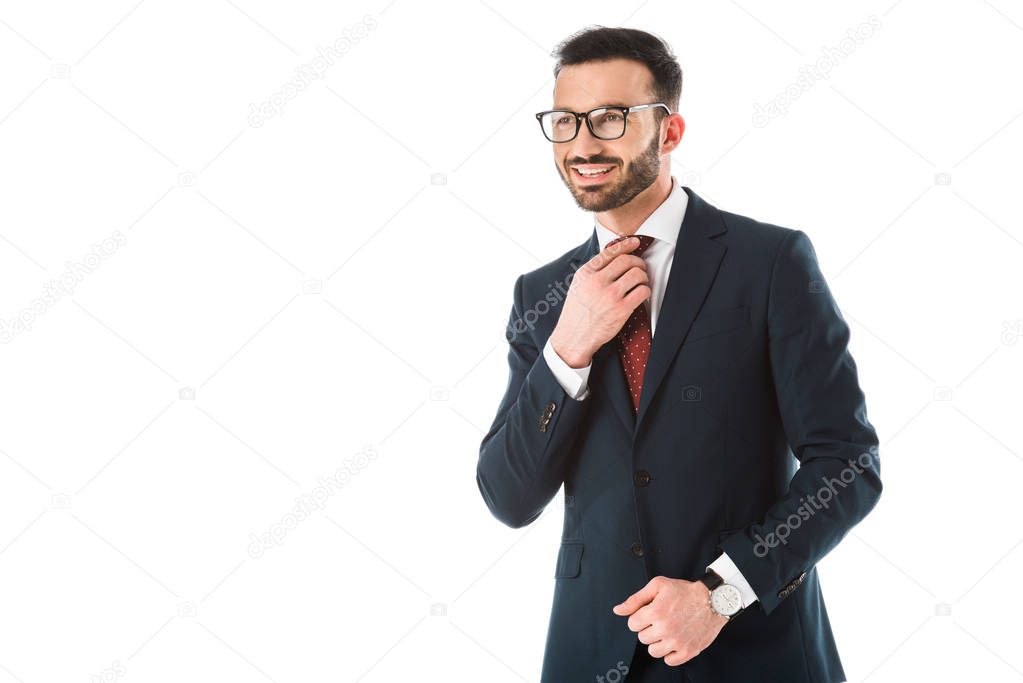 handsome smiling businessman touching tie and looking away isolated on white