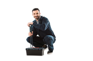 neşeli tesisatçı alet çantası yakınında otururken ayarlanabilir anahtarı tutarak ve beyaz izole kamera gülümseyerek
