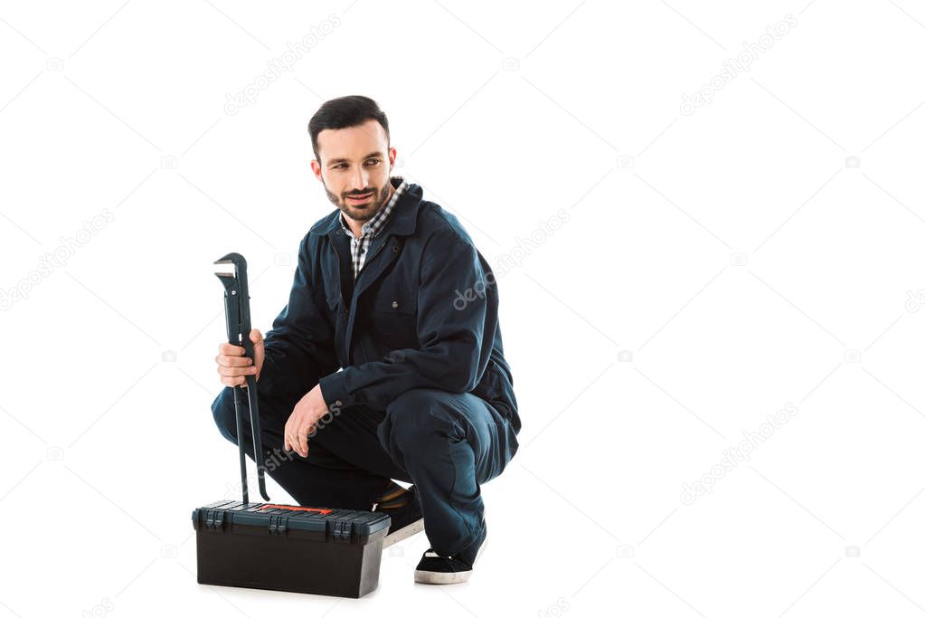 smiling workman holding adjustable wrench while sitting near toolbox isolated on white