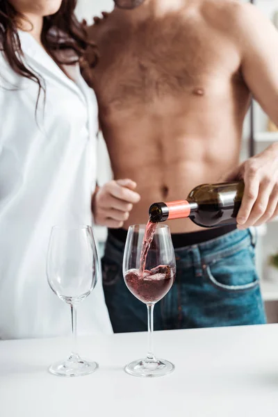 在厨房里女人附近的酒杯里倒红酒的无衬衫男子的裁剪视图 — 图库照片