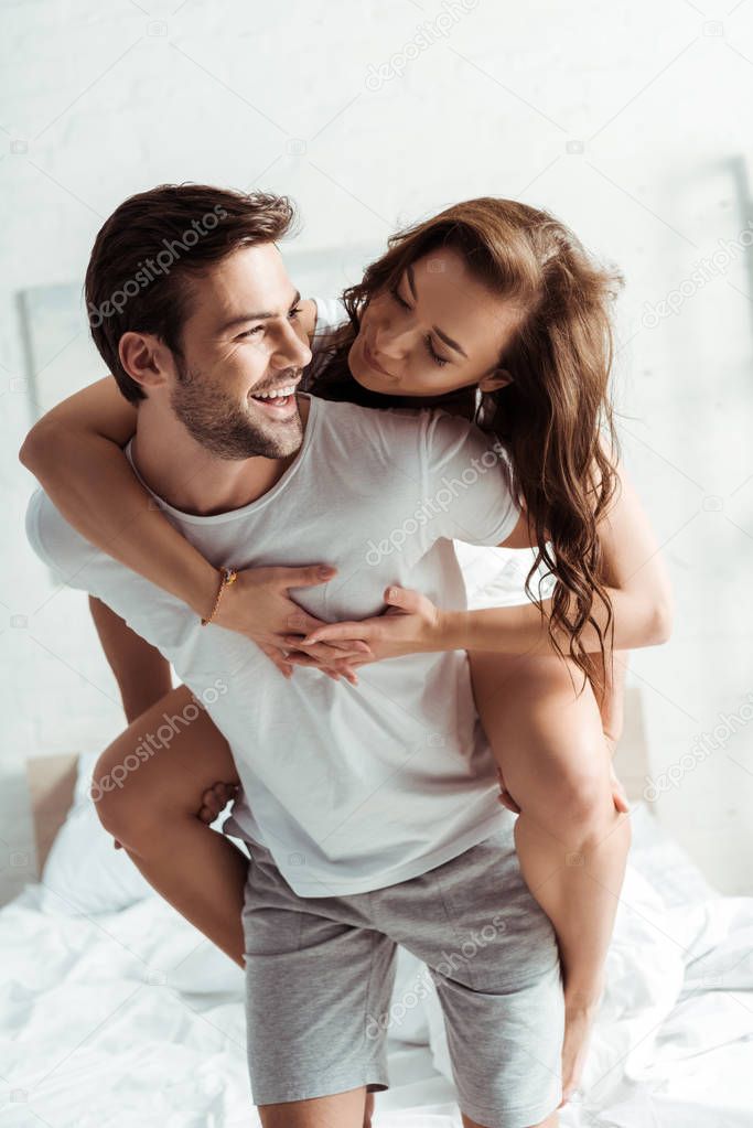 happy man piggybacking attractive girlfriend in bedroom 