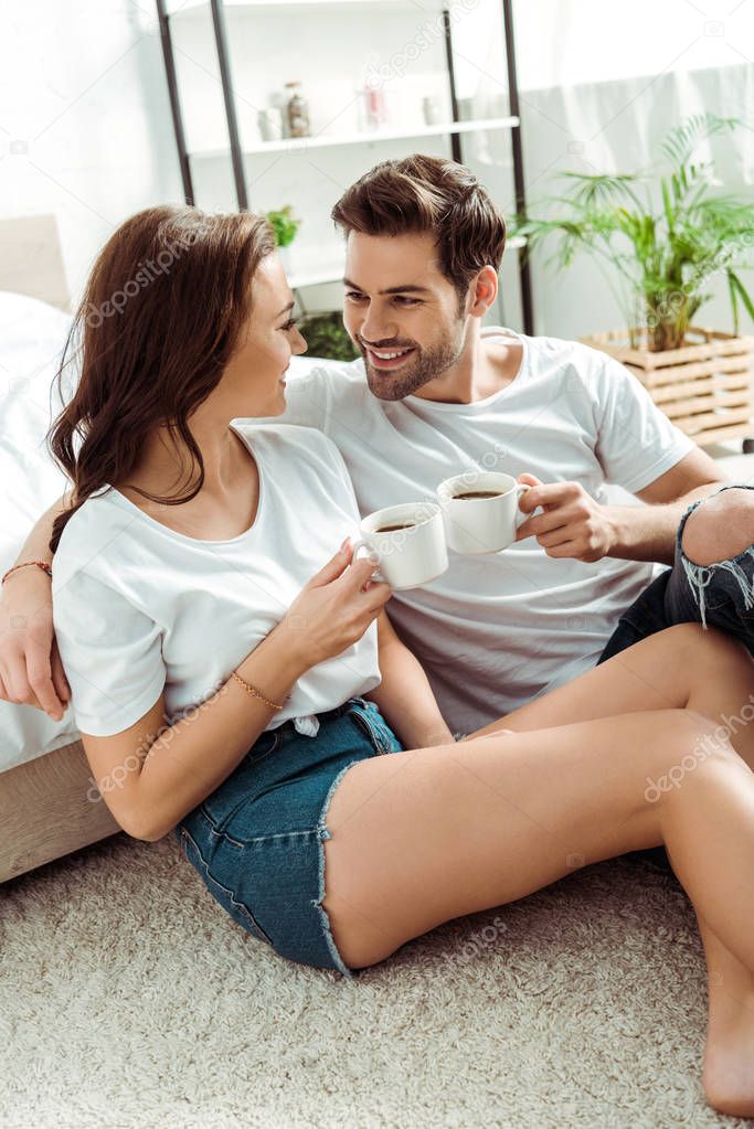 handsome man and cheerful woman holding cups of coffee in bedroom 