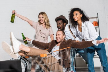 neşeli adam alışveriş sepeti sürme ve çok kültürlü arkadaşlar yakınında şişe tutarak 