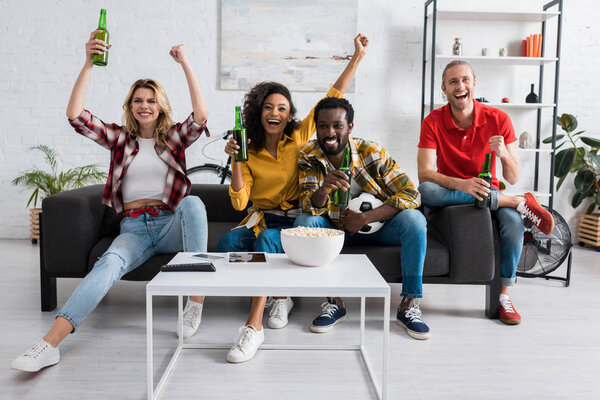 Smiling and positive multiethnic friends sitting on couch and drinking beer near table with bowl of tasty popcorn