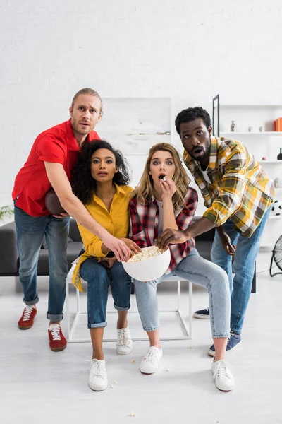 Genç çoklu etnik arkadaşlar maç izliyor ve oturma odasında lezzetli patlamış mısır yiyorlar.