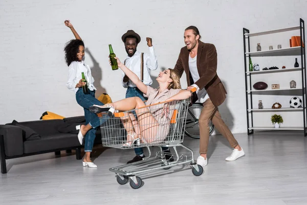 Happy Stylish Girl Riding Shopping Cart Multicultural Friends Holding Bottles — Stock Photo, Image