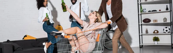 全景拍摄的快乐时尚女孩骑在购物车附近的多元文化的朋友拿着啤酒瓶 — 图库照片