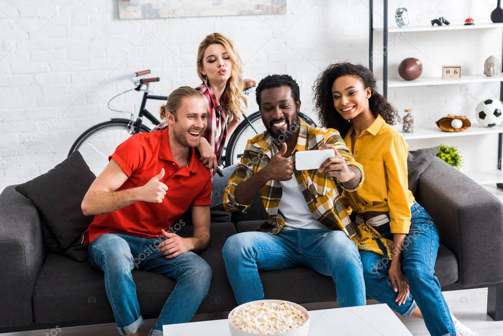 Excited happy multicultural men and women sitting on couch and taking selfie in living room