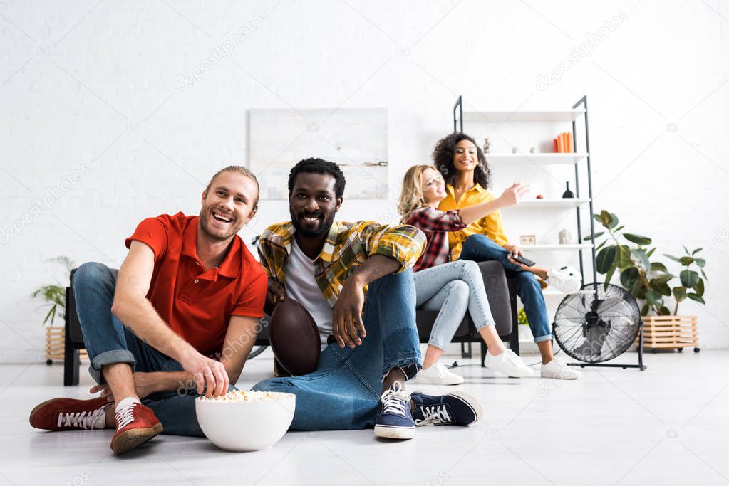 Two multicultural men watching match and eating popcorn while two women taking selfie