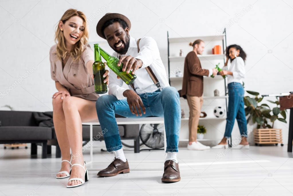 selective focus of happy girl laughing and clinking bottles of beer with cheerful african american man 