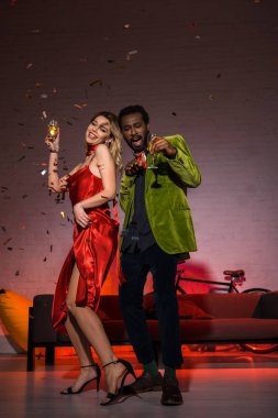 kırmızı elbiseli sarışın kadın ile ayakta ve şampanya cam tutan mutlu afrikalı-amerikalı adam düşük açı görünümü 