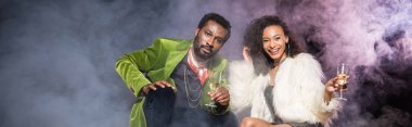 duman ile siyah konfeti yakınında otururken şampanya bardakları tutan afrikalı Amerikalı adam ve kadın panoramik çekim 