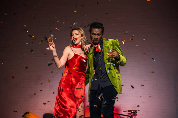 с низким углом зрения на африканского американца, стоящего с блондинкой в красном платье и держащего бокал шампанского
 