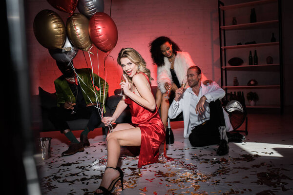 избирательный фокус веселой блондинки в красном платье с воздушными шарами и бокалом шампанского рядом с мультикультурными друзьями
 