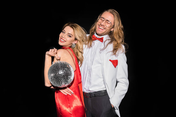 cheerful blonde girl holding shiny disco ball and smiling near man in glasses isolated on black 