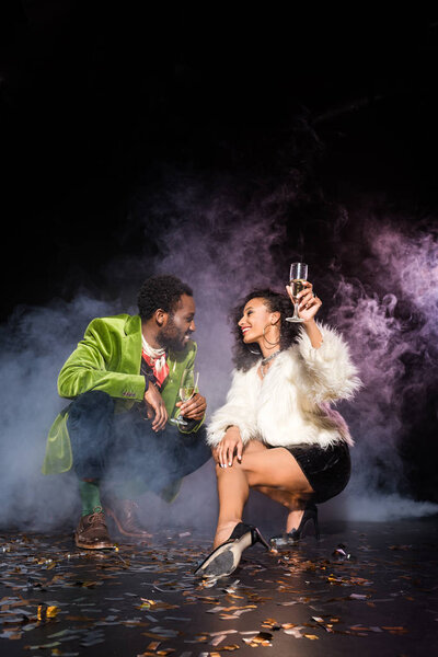 african american man and woman holding champagne glasses while looking at each other near confetti on black with smoke 
