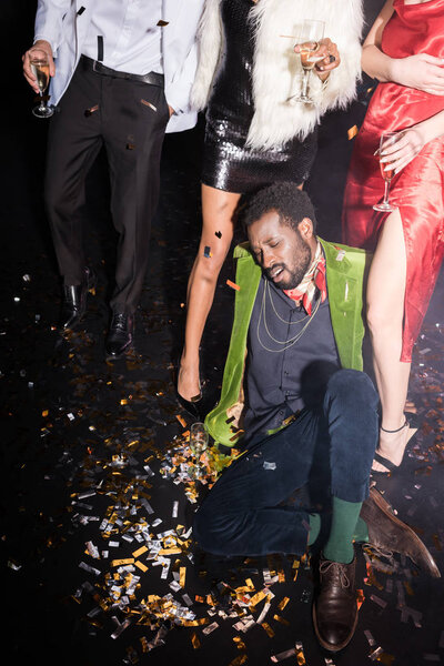 handsome and drunk african american man lying on floor with shiny confetti near friends on black 