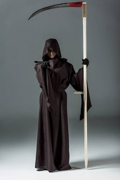 full length view of woman in death costume holding scythe and pointing with finger on grey