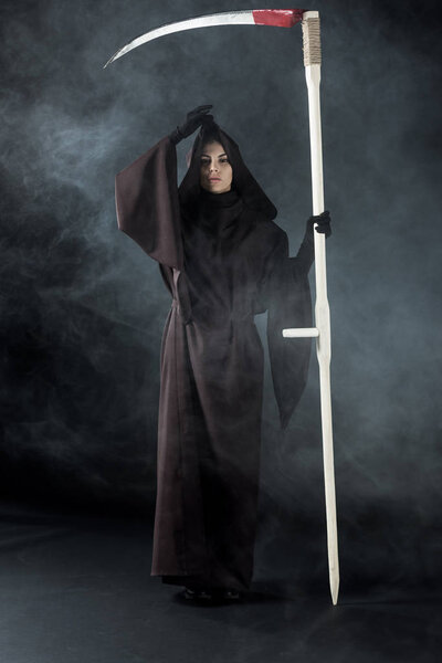full length view of woman in death costume holding scythe on black with smoke
