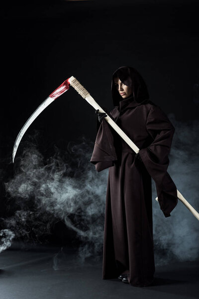 full length view of woman in death costume holding scythe on black with smoke