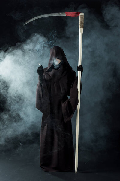 full length view of woman in death costume holding scythe and smoking cigarette on black